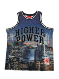 A blue mesh basketball jersey with 'HIGHER POWER' printed in black arched text across the front, featuring a city skyline graphic and a red Mitchell & Ness neck label.